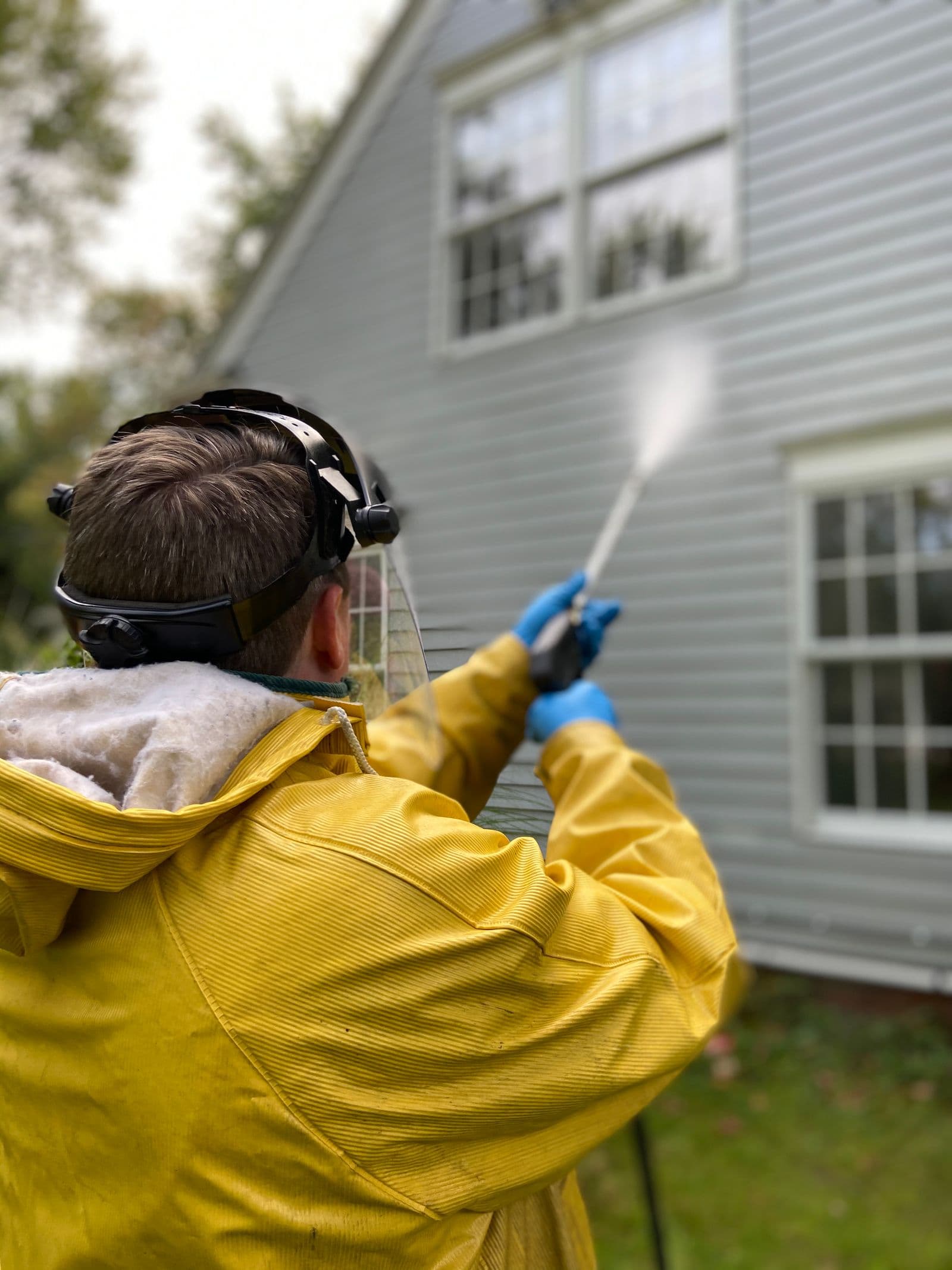 Pressure-washing technician in protective gear soft-washing the vinyl siding of a two-story home